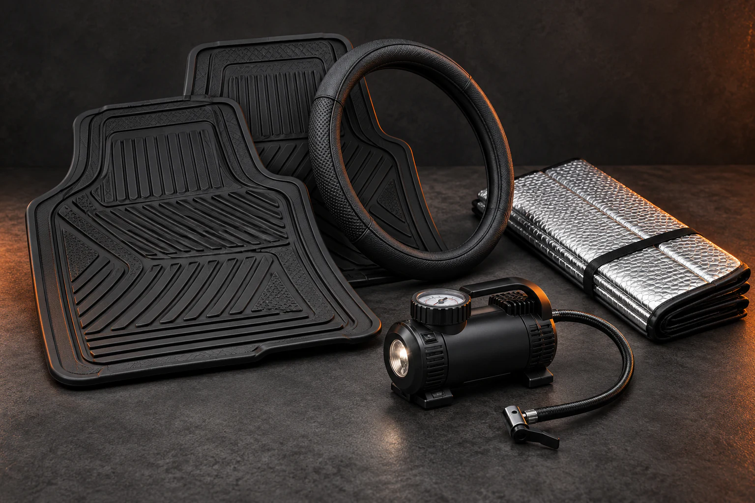 Illustration of unbranded rubber car mats, steering-wheel cover, reflective sunshade and compact tyre inflator on a charcoal surface with orange lighting